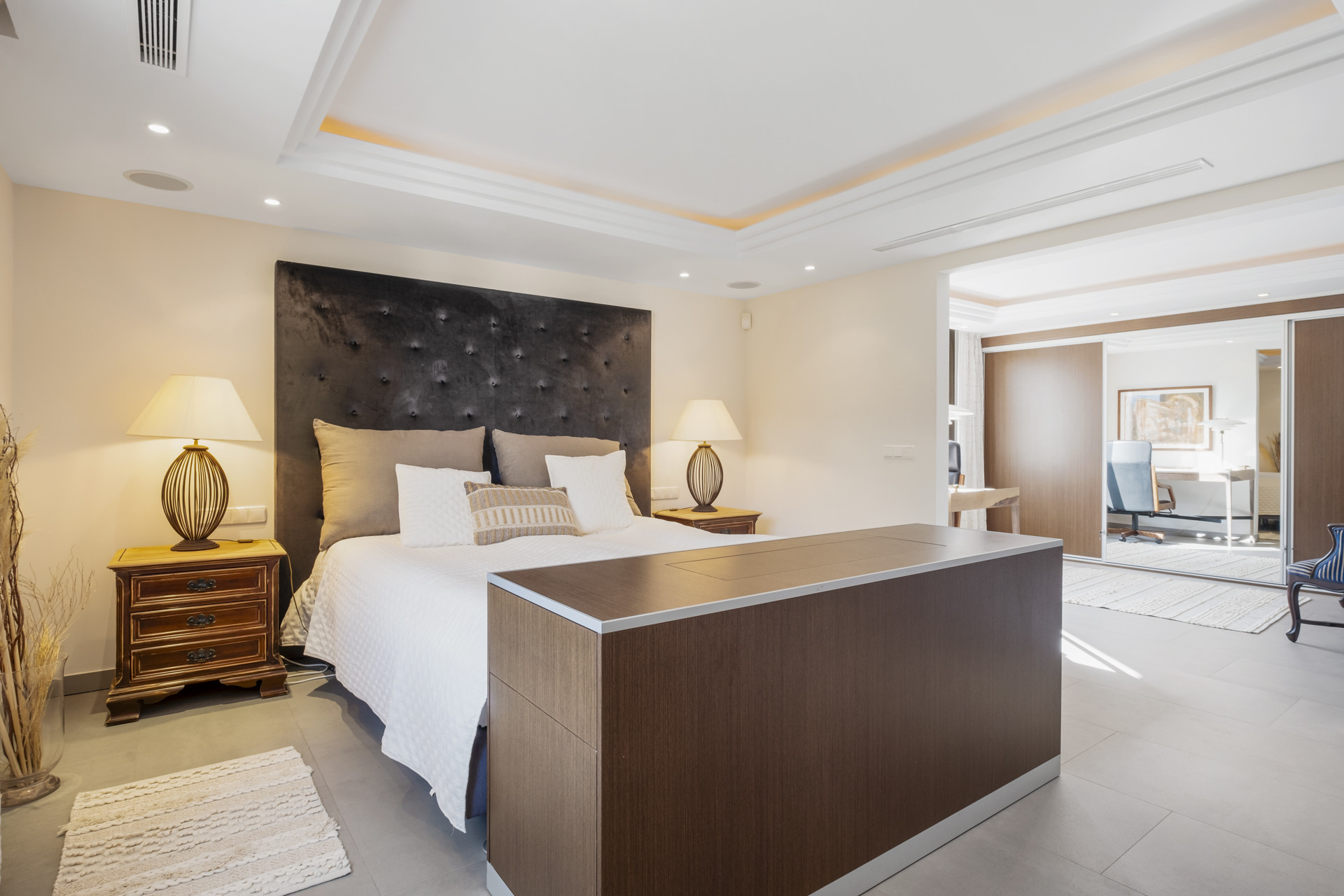 Cozy bedroom featuring a large bed and nightstands, with a workspace visible in the background.