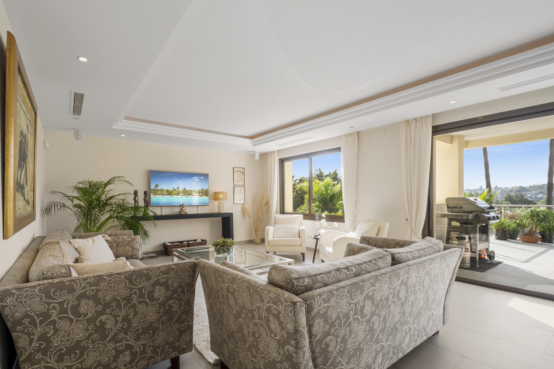 Bright living room with comfortable seating and a TV, opening to a balcony with grill.