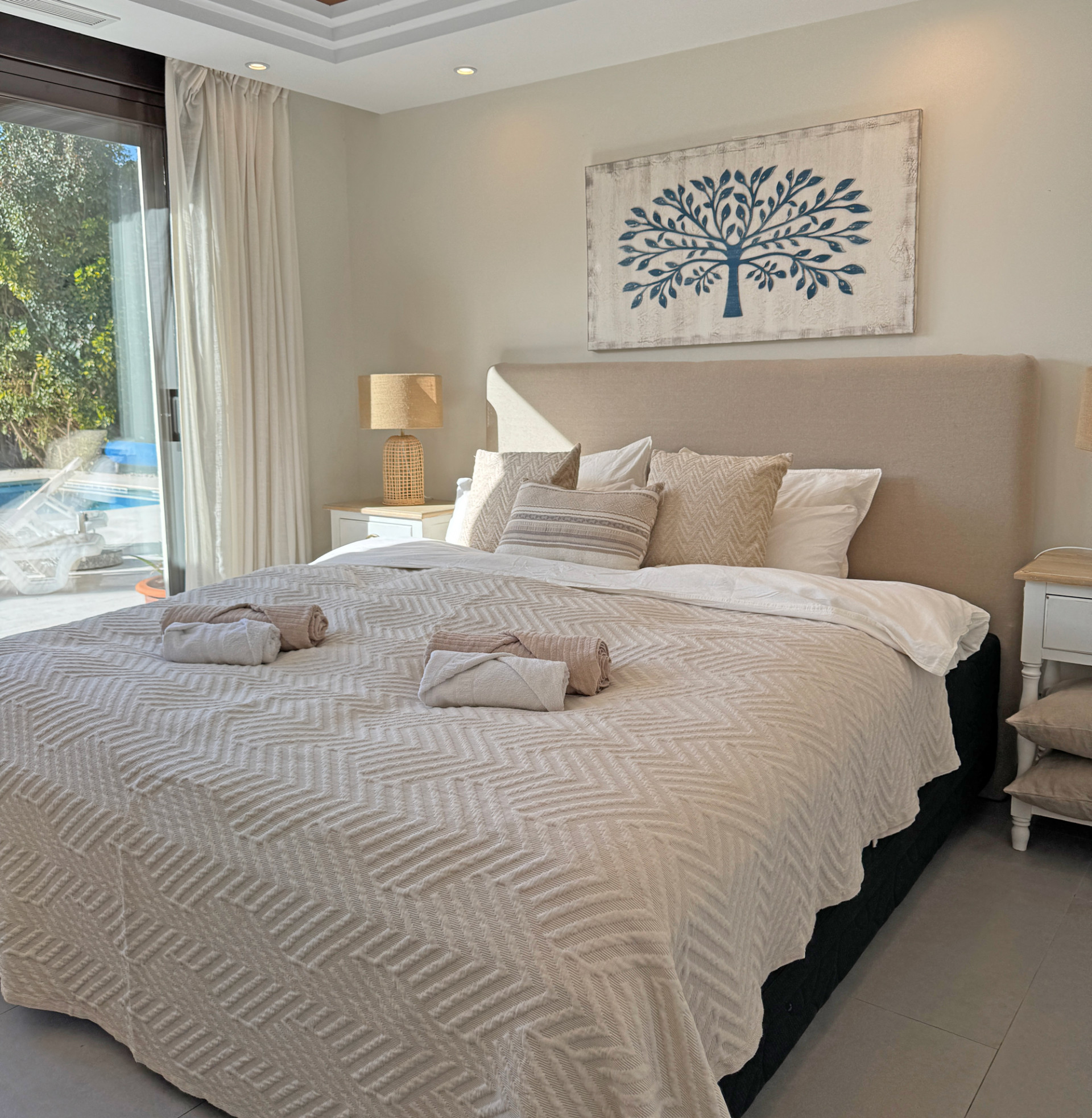 Warm bedroom featuring neatly made bed, decorative pillows, and visible pool outside through the glass door.
