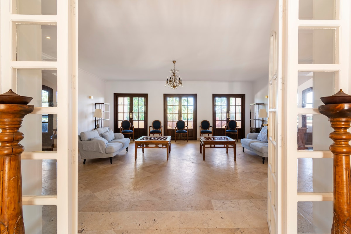 Living room view showcasing a chandelier, wooden furniture, and large windows.