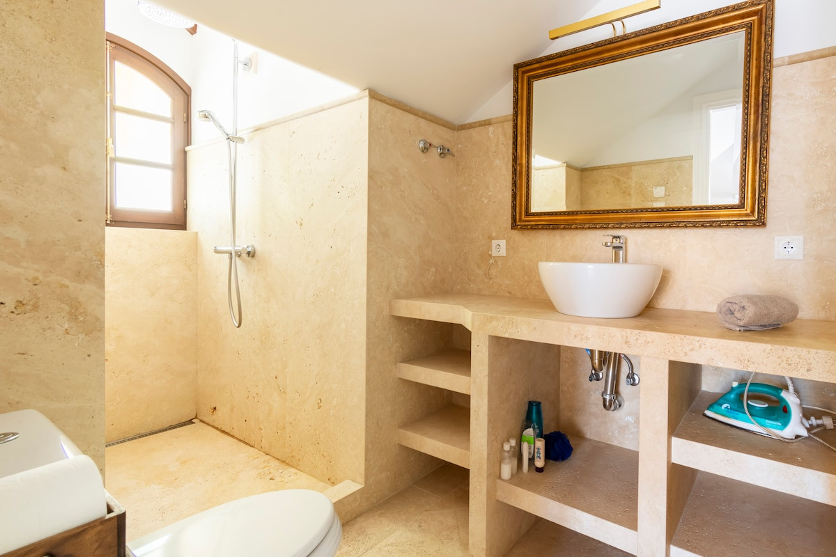 Bright bathroom with beige marble surfaces and a shower area with a window.