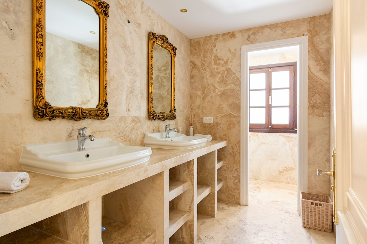 Bathroom with dual sinks, ornate mirrors, and beige stone walls.