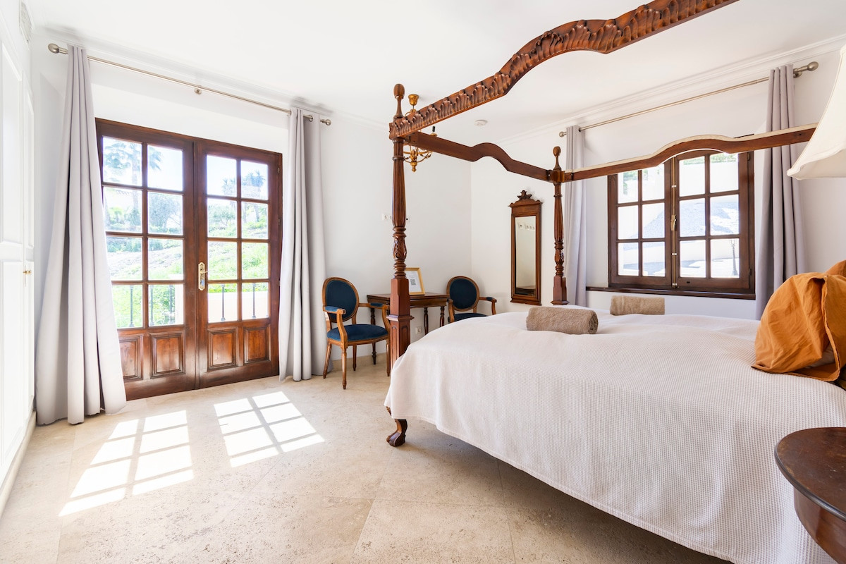 Bright bedroom with a four-poster bed, chairs, and French doors to the outside.