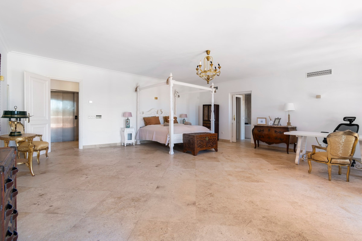 Large bedroom featuring a four-poster bed, a chic chandelier, and elegant furnishings with natural light.