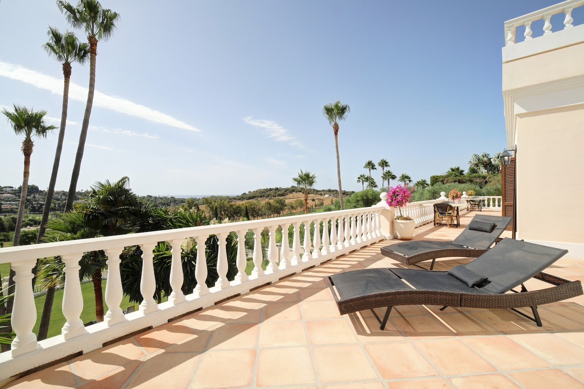 Outdoor terrace with sun loungers and a view of the surrounding landscape and palm trees.