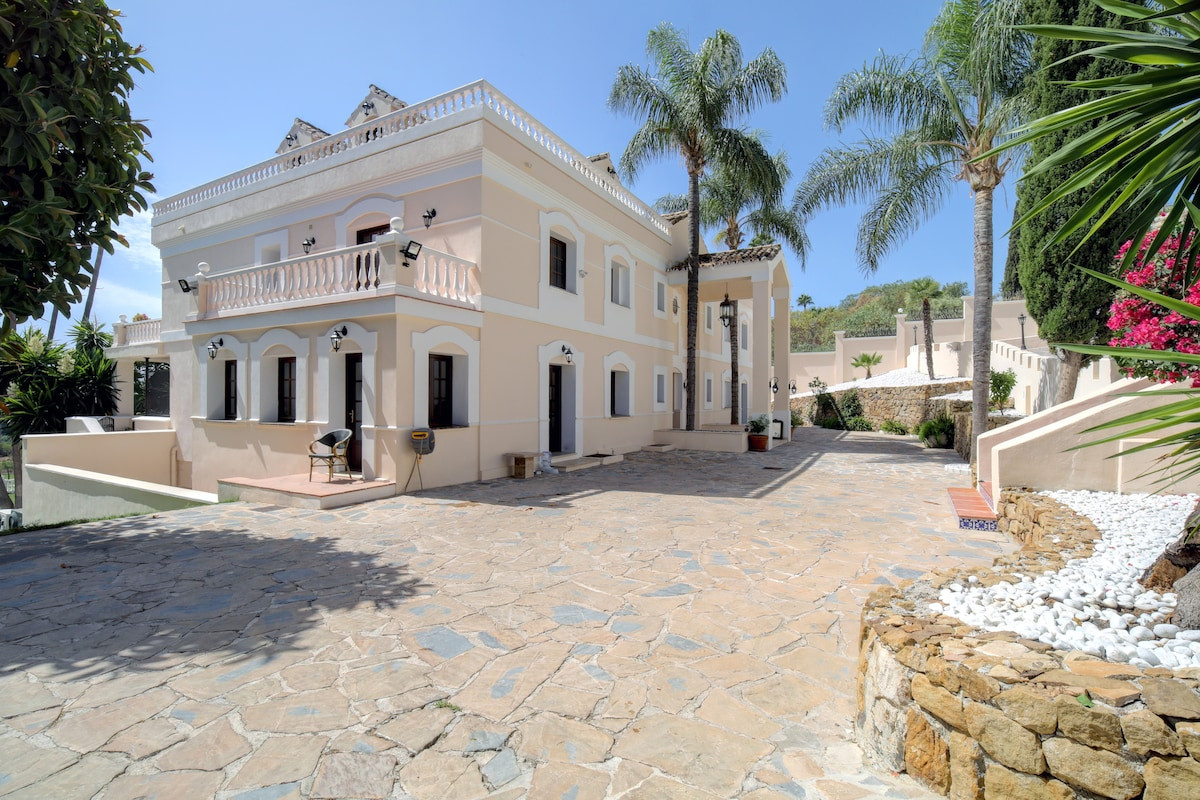 Exterior view of a house with a stone paved area and palm trees, showcasing balconies and light colored walls.