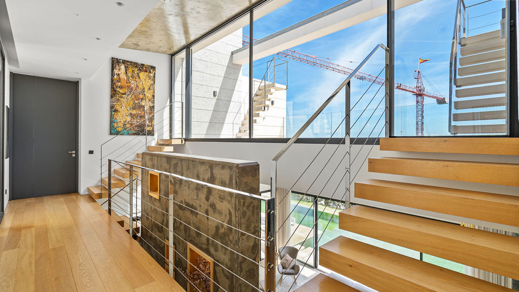 Stairway with wooden steps and glass railing, adorned with artwork and large windows providing ample sunlight.