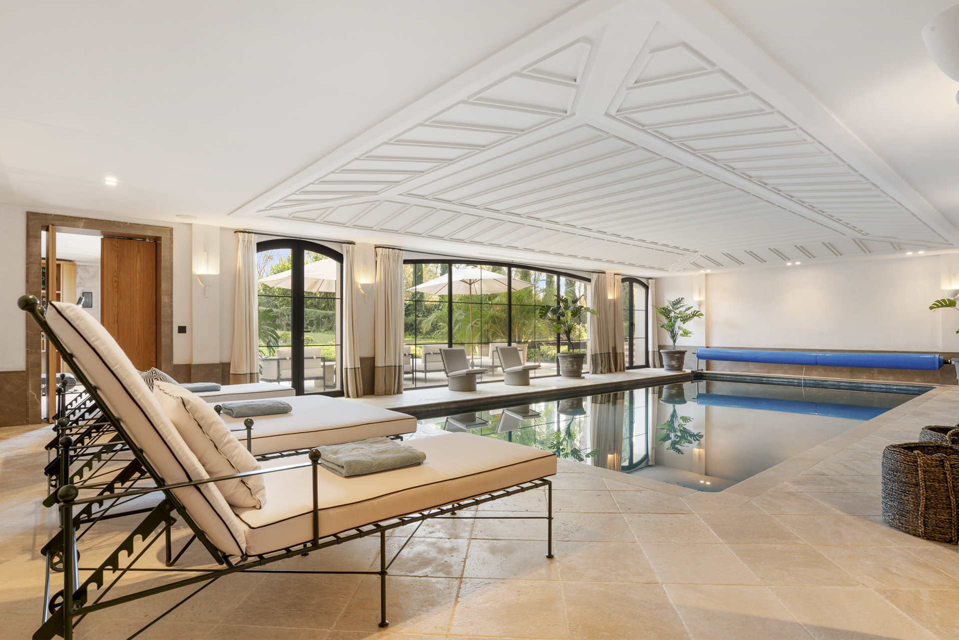 Indoor pool area with lounge chairs, large windows overlooking the outdoor patio and greenery.