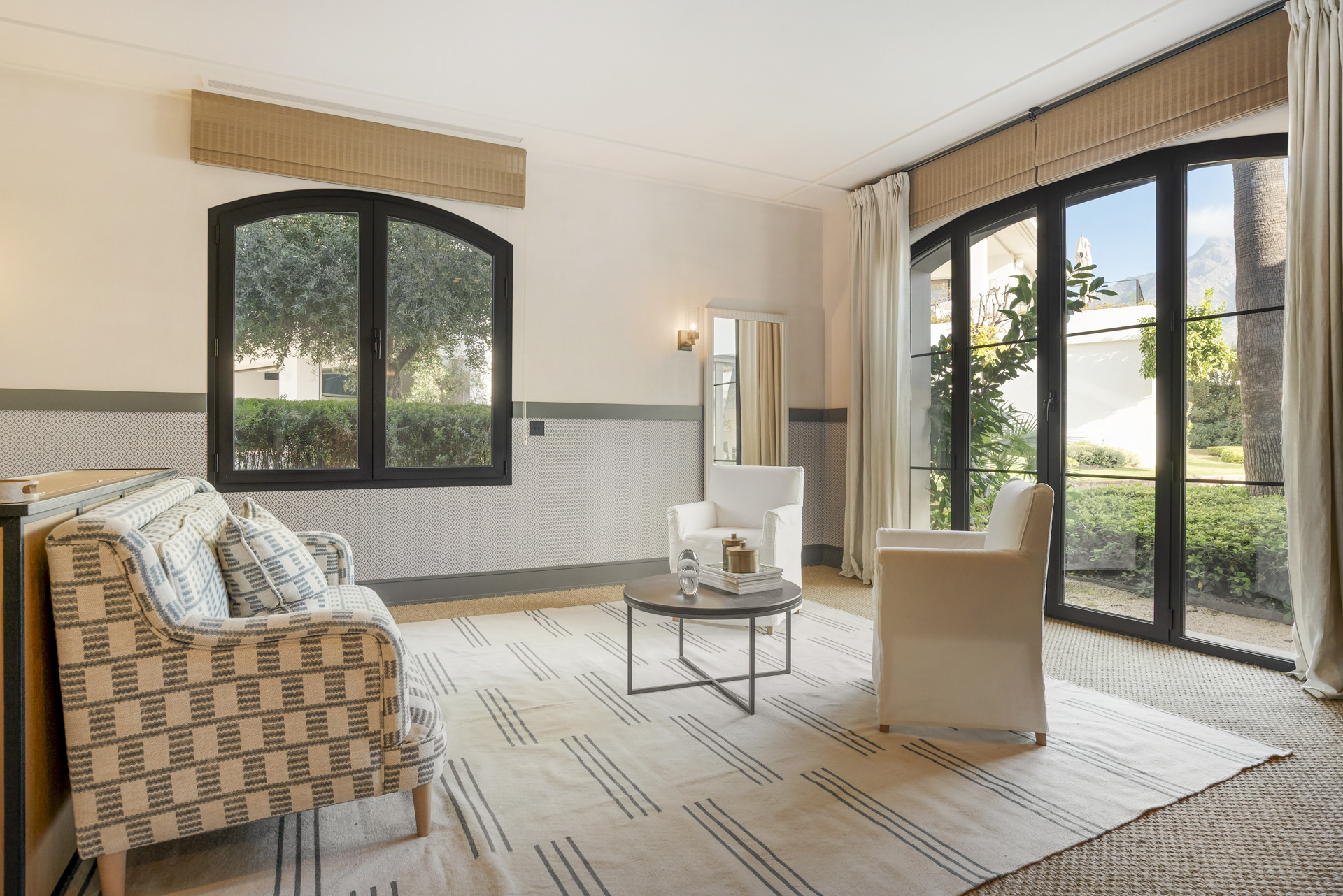 Living room with patterned chairs, a table, and large windows overlooking greenery.