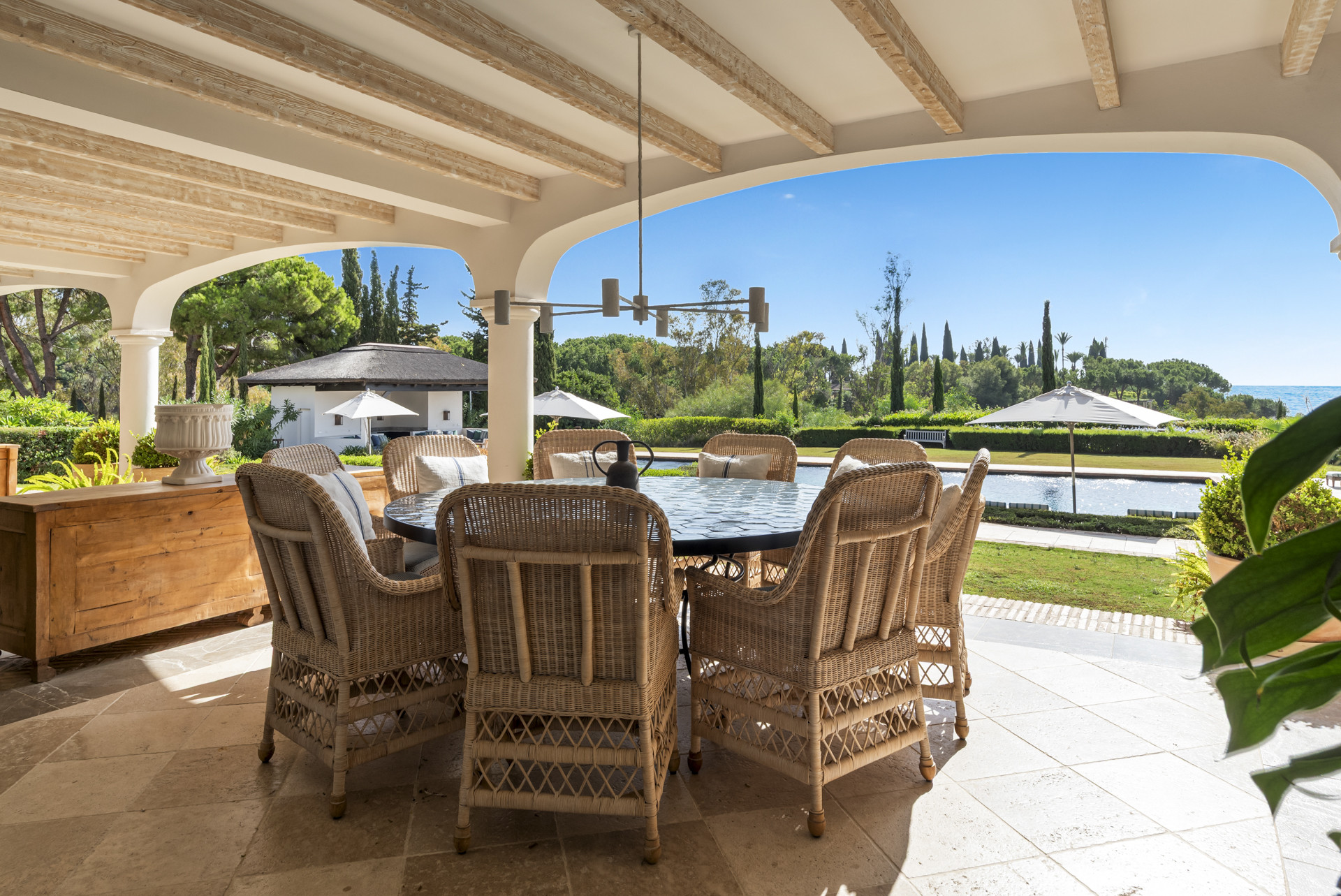 Covered terrace with a large dining table and wicker chairs overlooking the garden and pool area.