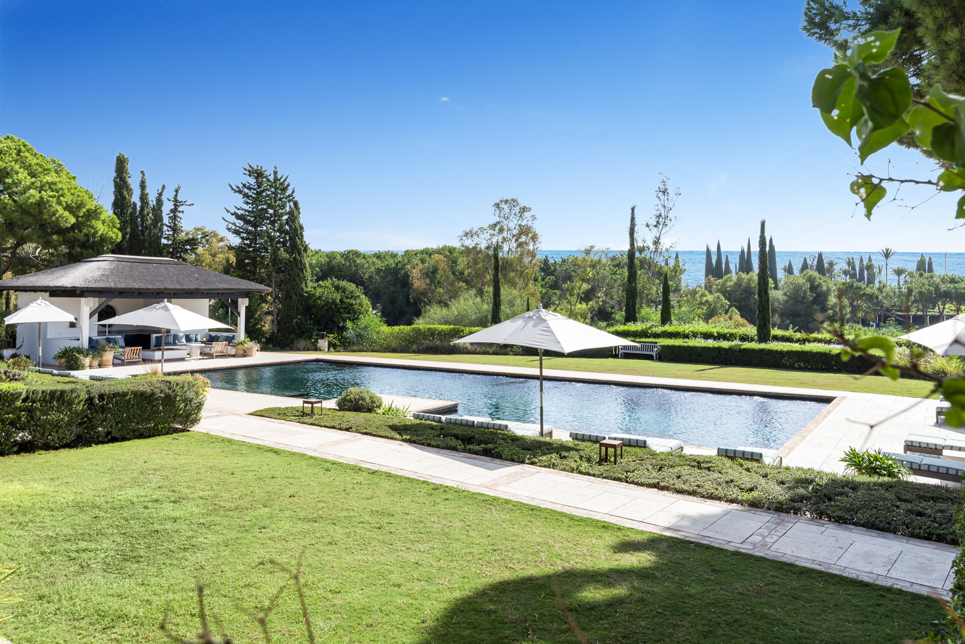 Outdoor pool area surrounded by gardens, featuring shaded seating and a view of lush greenery and sky.