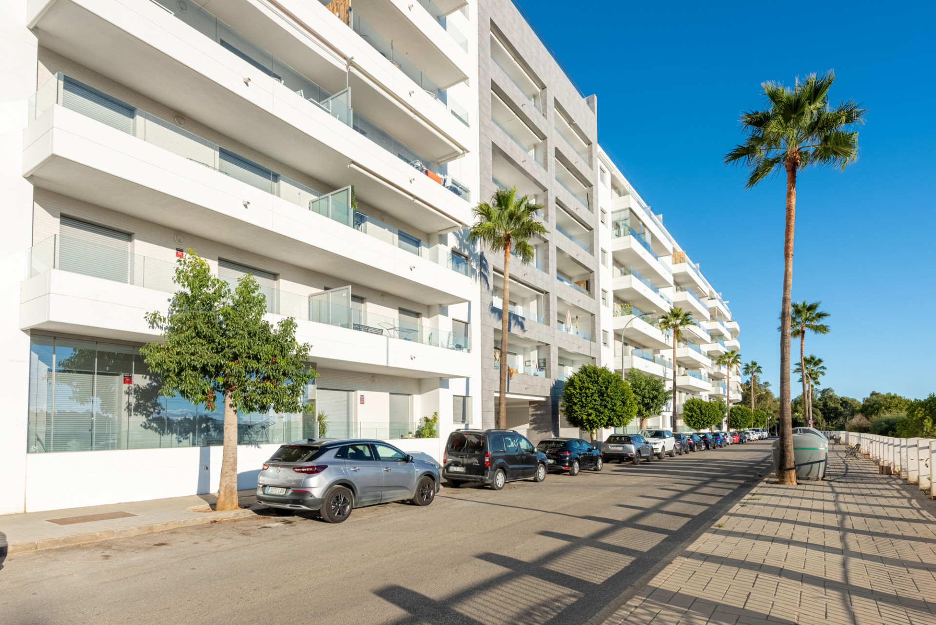 Exterior view of a modern apartment building with balconies, palm trees, and parked cars on the street.