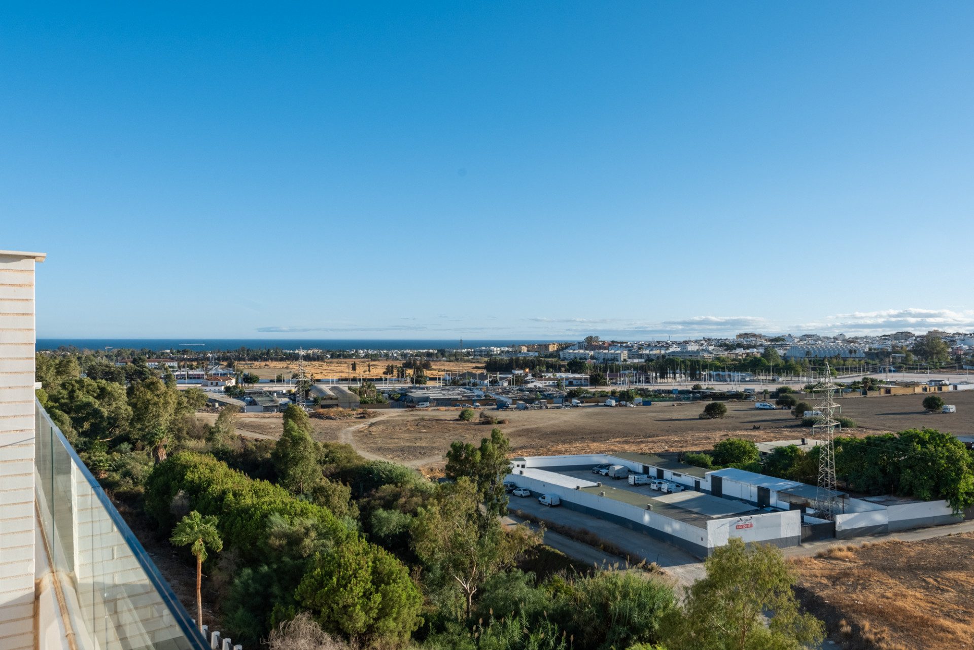 Wide view of the coastline and city from a high balcony with a glass railing and green trees.