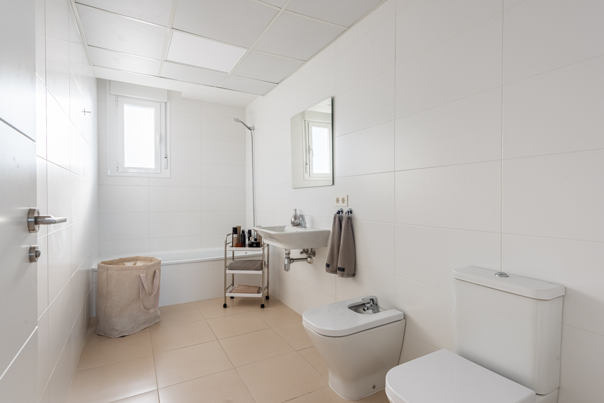 White tiled bathroom with a bathtub, bidet, toilet, and sink. A small window is above the tub.