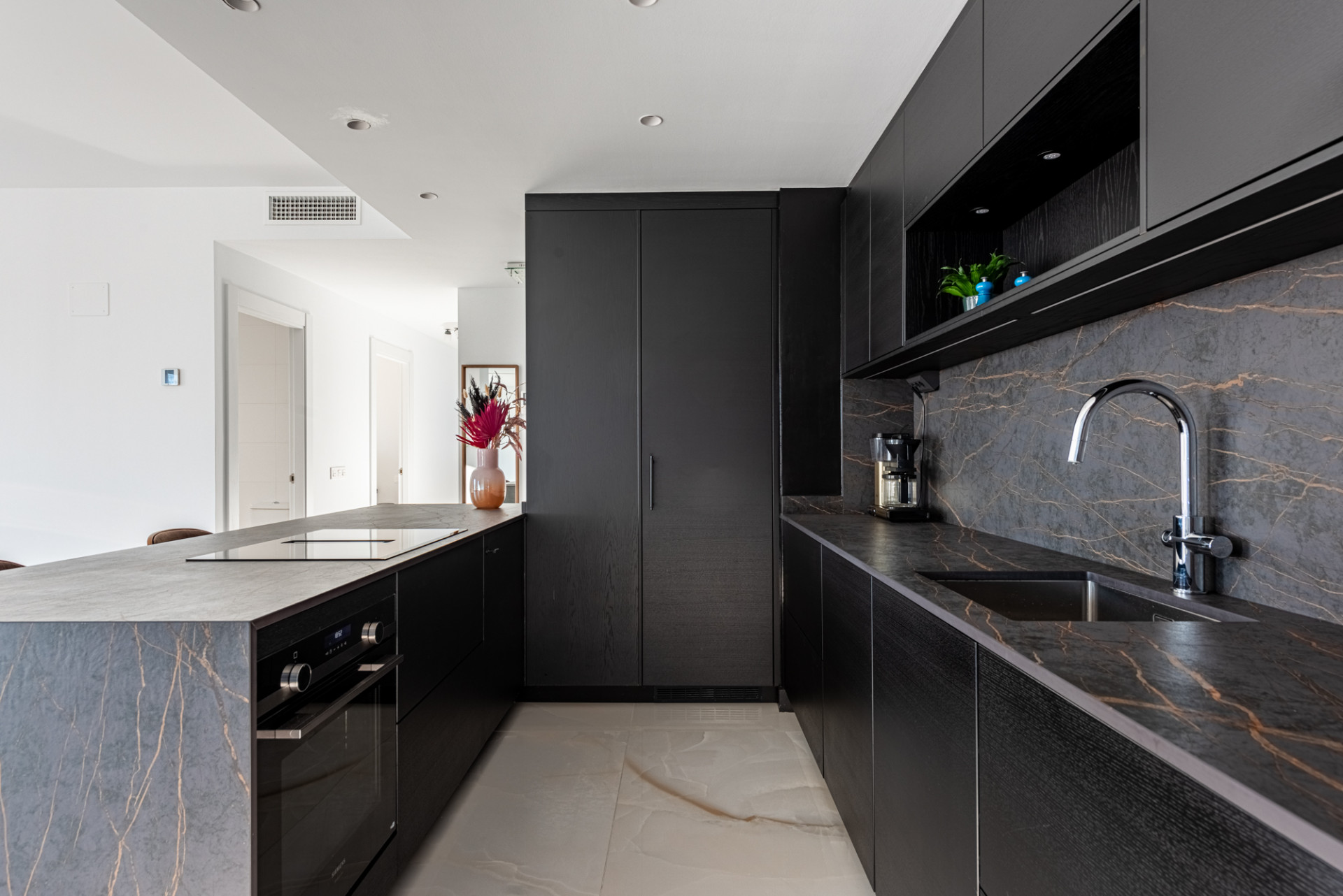 Dark kitchen with black cabinets, gray marble surfaces, and built-in oven and induction hob.