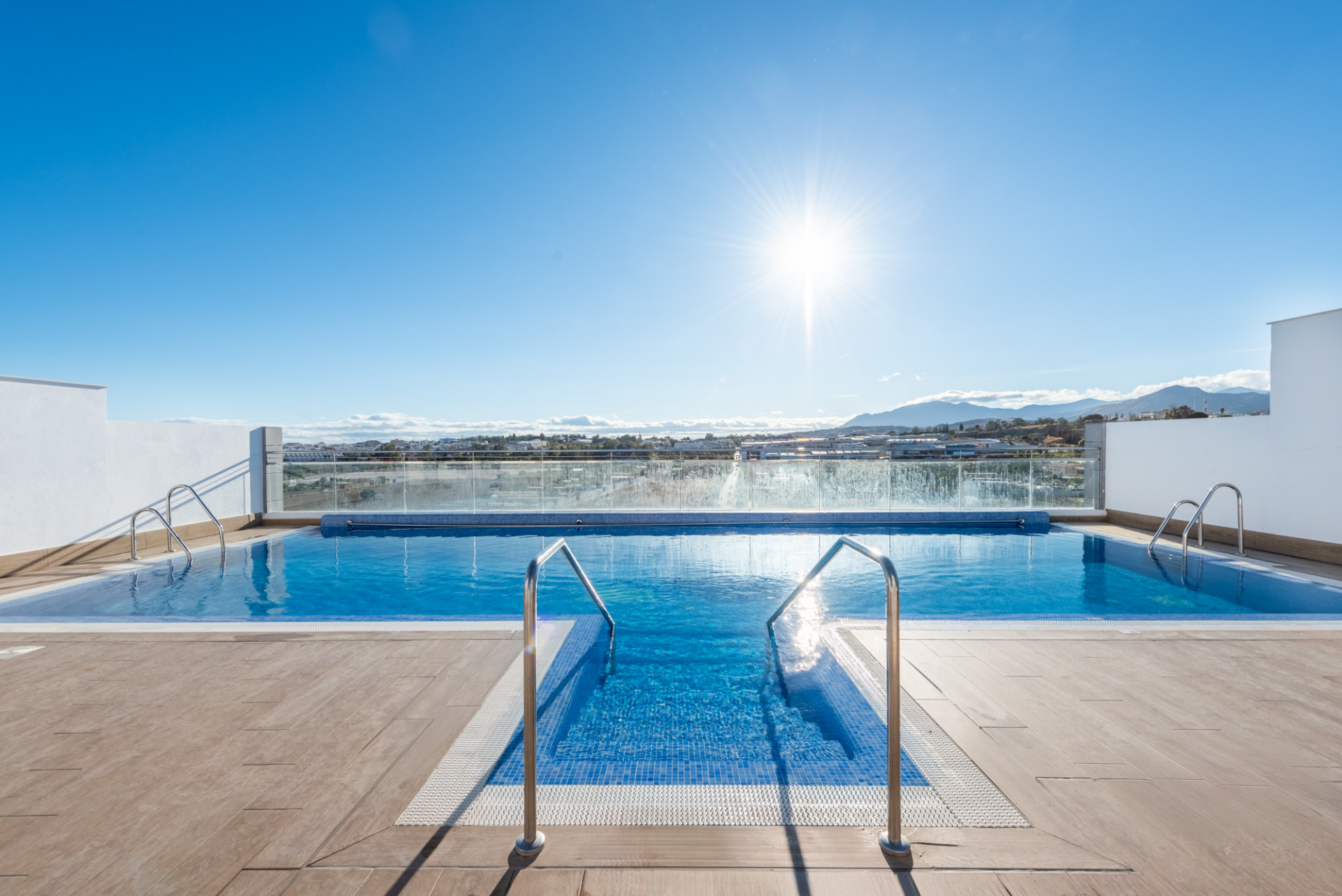 Rooftop infinity pool with clear blue water and sunny sky, mountains in the distance.