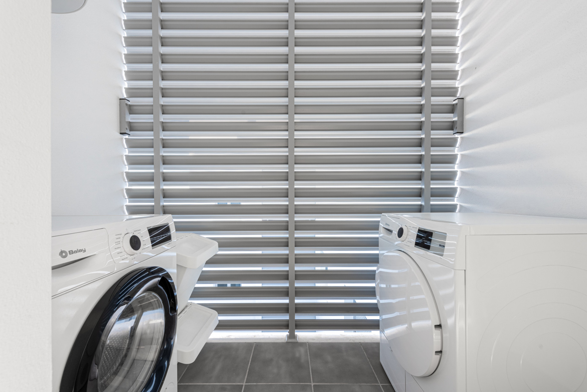 Laundry area with a front-loading washing machine and a dryer, facing slatted window blinds.