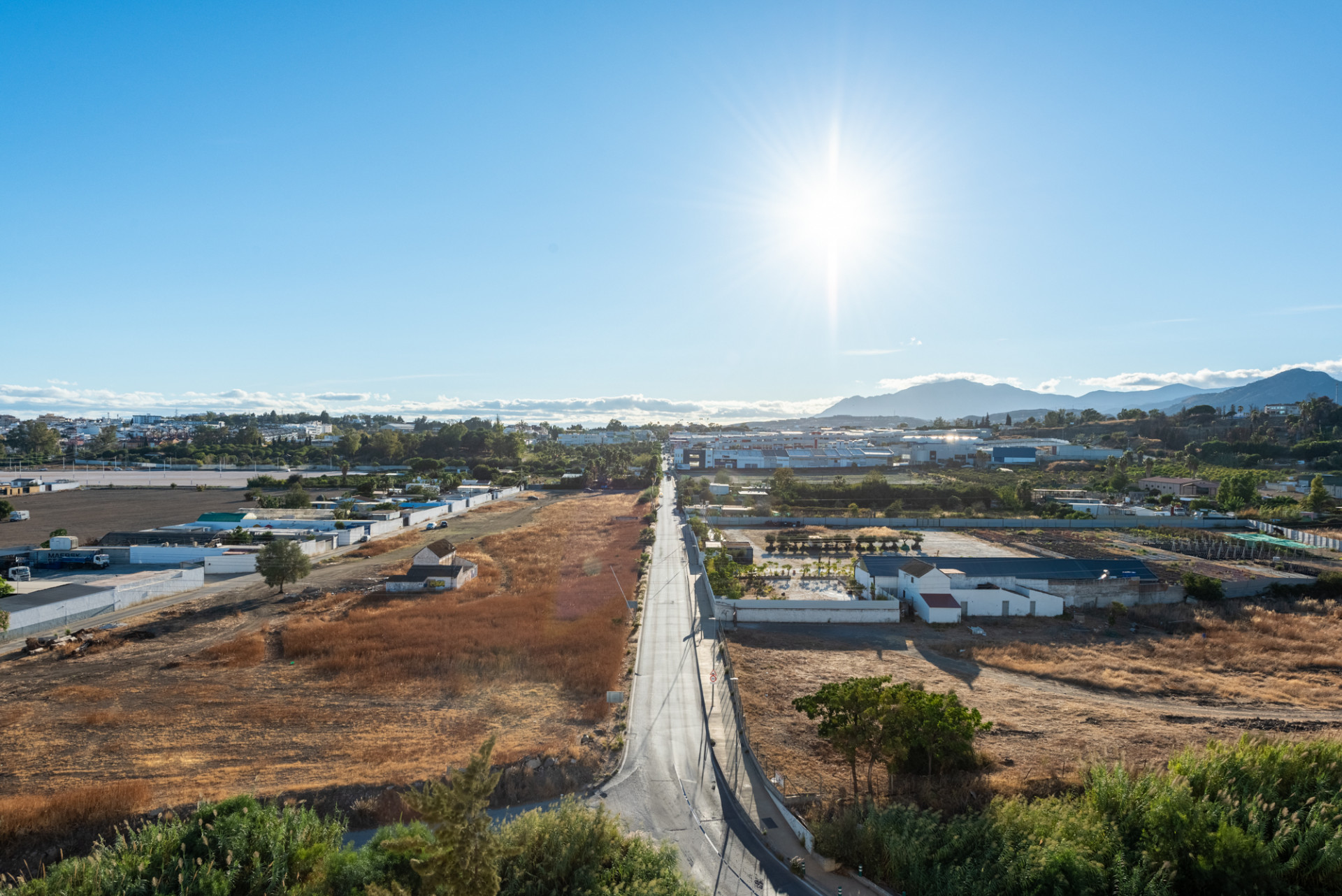 Aerial view of a long road through a rural area, with distant mountains and a bright sun in the sky.