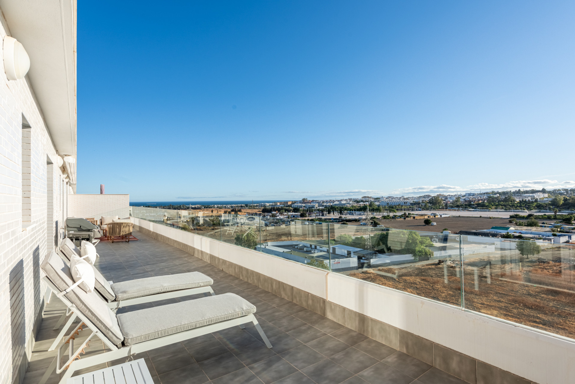 Rooftop terrace with two lounge chairs, glass railing, and a view of the distant city and sea.