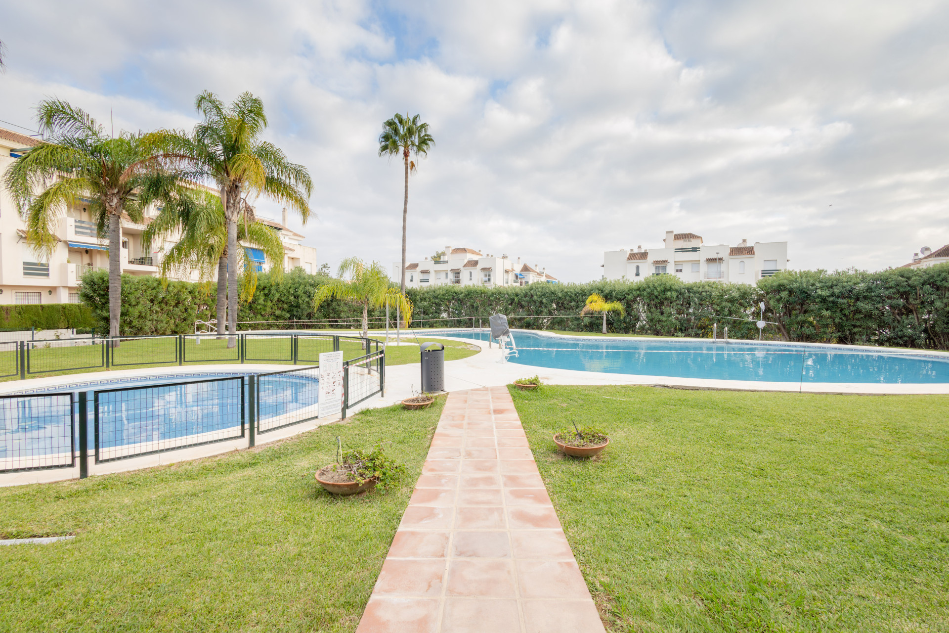 Outdoor communal area with two swimming pools, palm trees, and residential buildings under a cloudy sky.