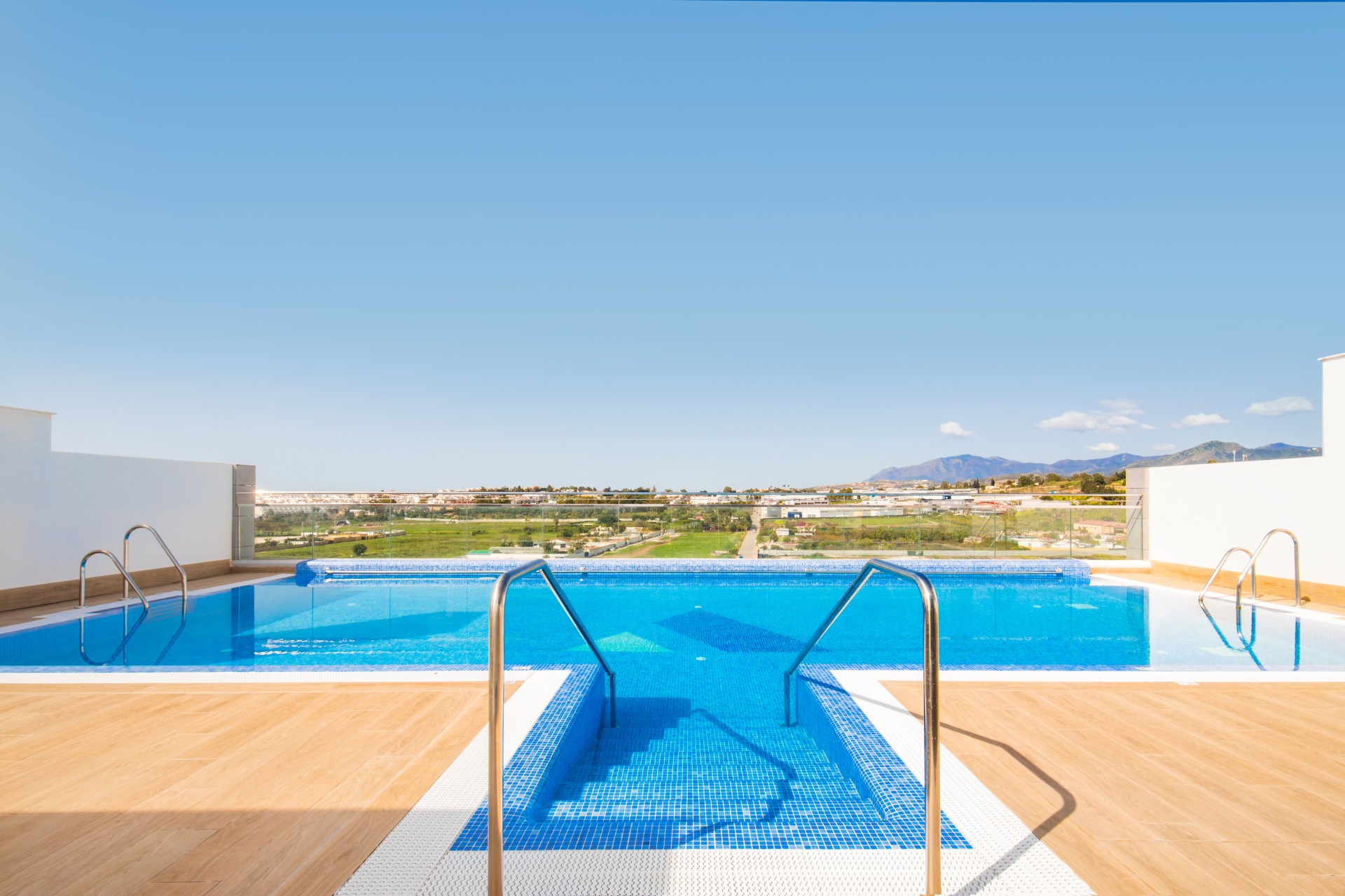 Infinity pool with stairs, surrounded by wood decking, overlooking green landscape.