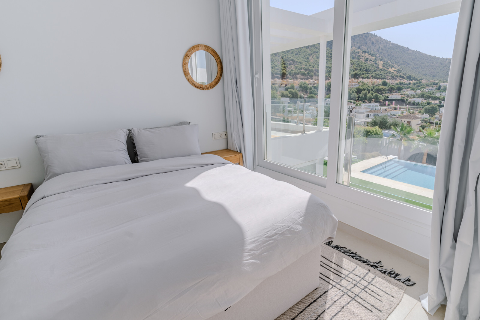 Bedroom with large windows, light bedding, and a view of the pool and mountains.