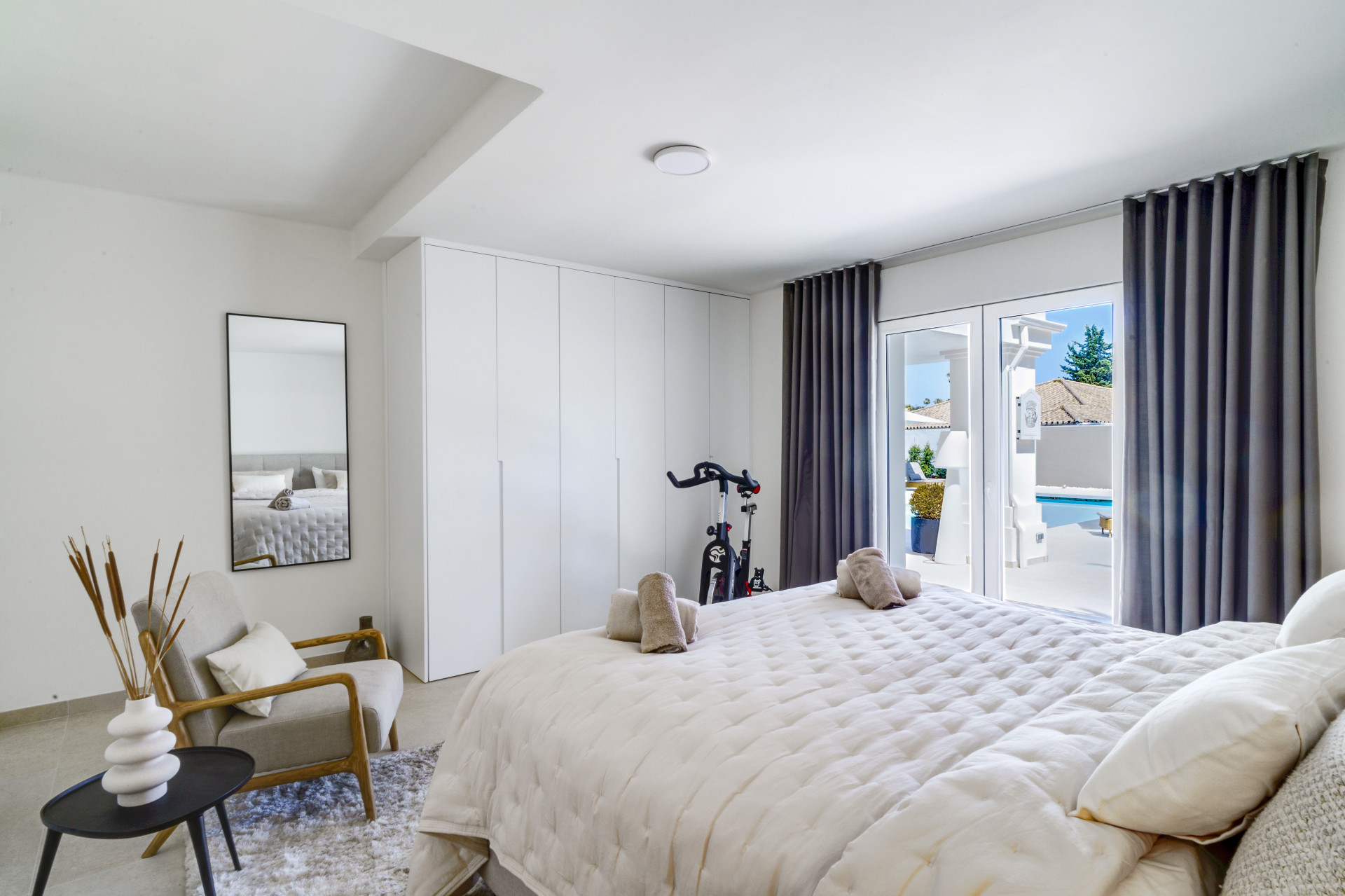 Bright bedroom with large window and view of the pool area, features a bike and minimalist decor.