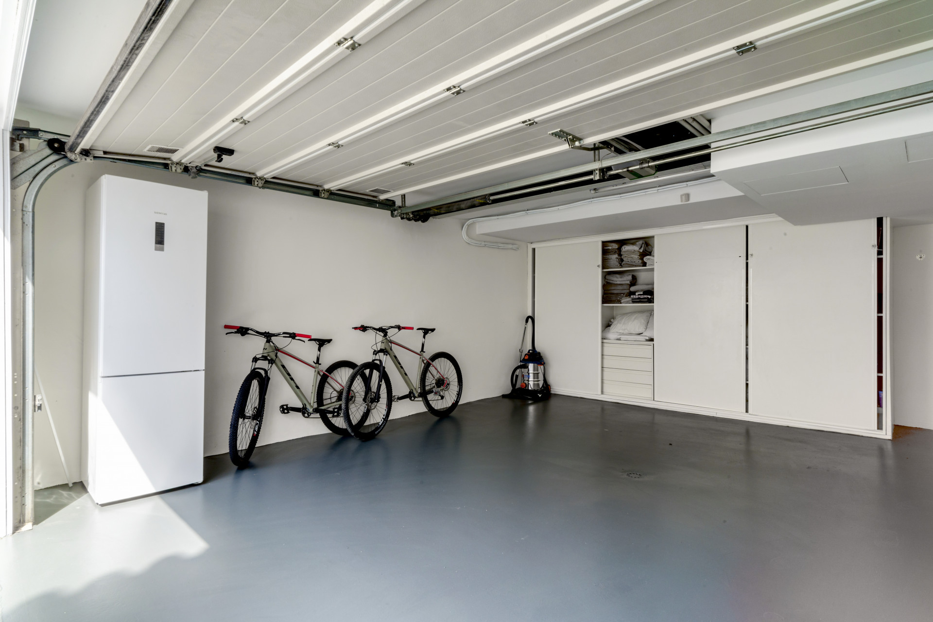 Garage containing two bicycles, a refrigerator, and shelving units against a white wall.
