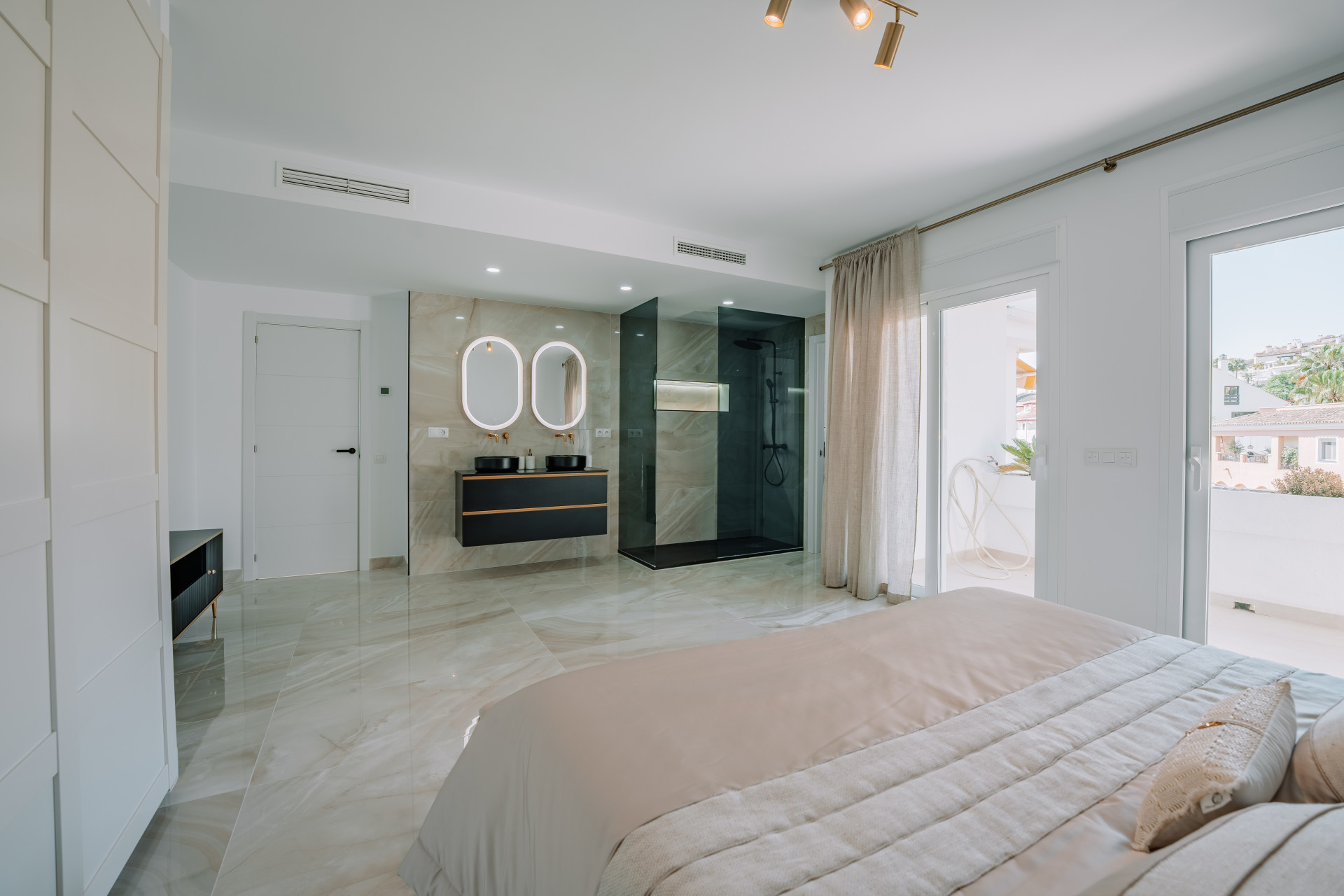 Bedroom with a bed on the right. A double vanity with backlit mirrors and a walk-in shower are integrated into the room.