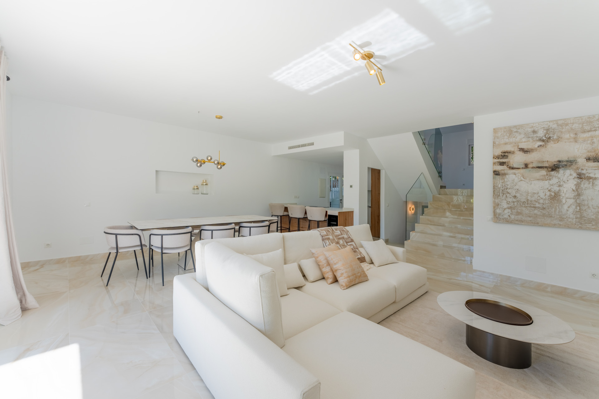 Bright open-plan living room with white sectional sofa, dining area, and marble staircase.