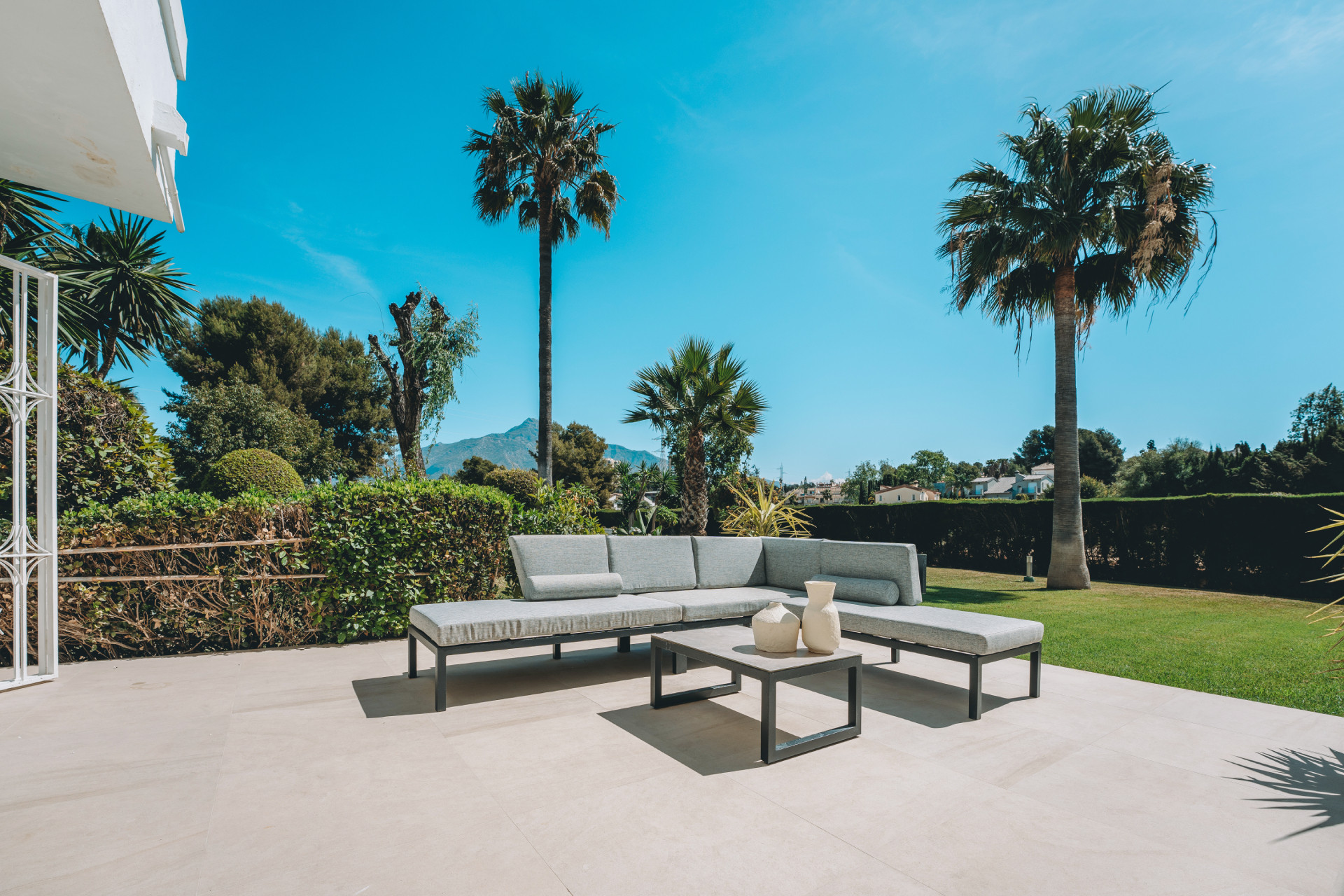 Outdoor patio with a large grey sectional sofa, coffee table, and palm trees under a blue sky.