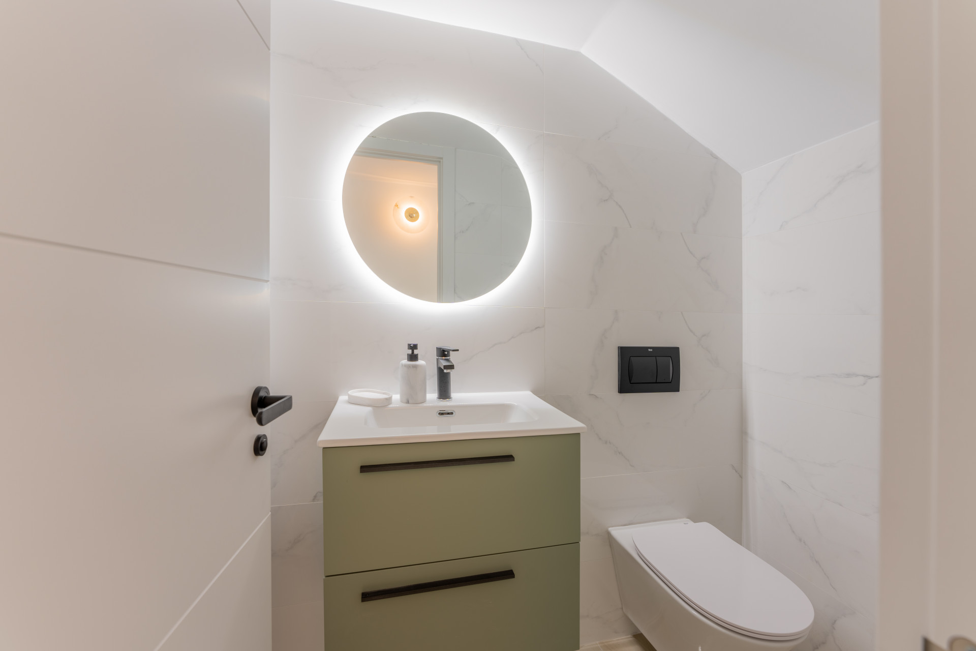 Modern bathroom with white marble-effect tiles, a green vanity, and a backlit circular mirror.