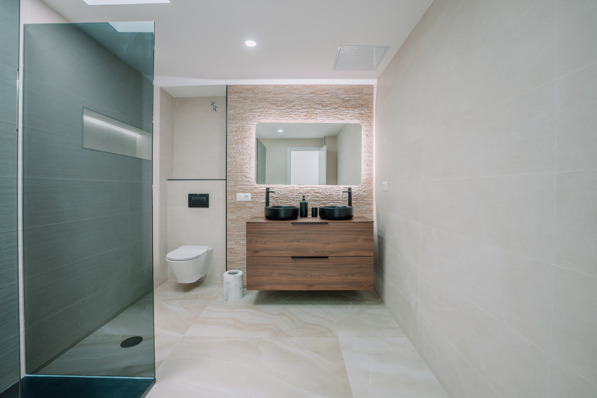 Bathroom with a walk-in shower on the left, a wall-mounted toilet, and a vanity with two black vessel sinks.