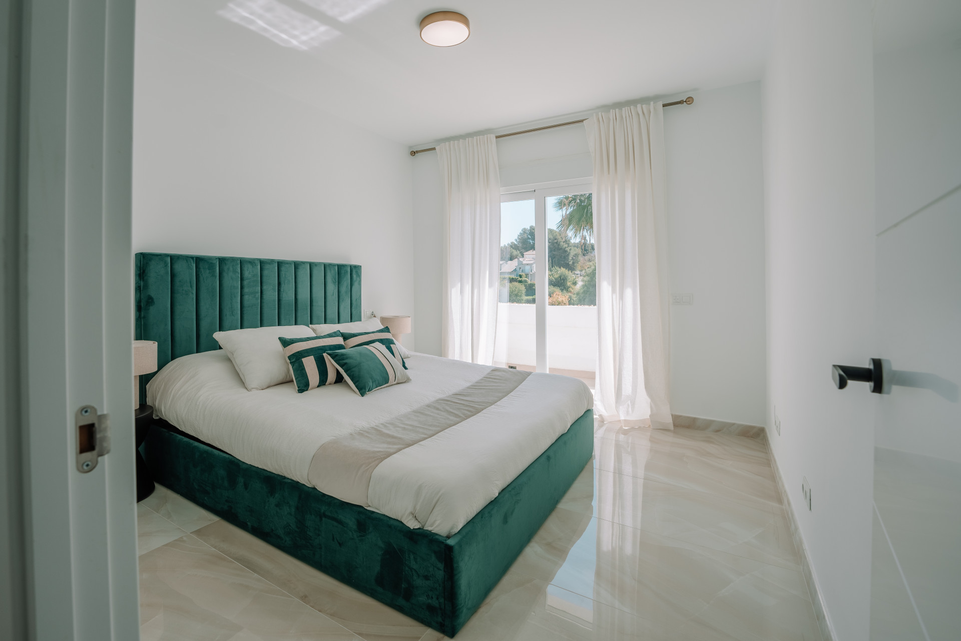 Bright bedroom with a large green velvet bed, white walls, and glass doors leading to a balcony.