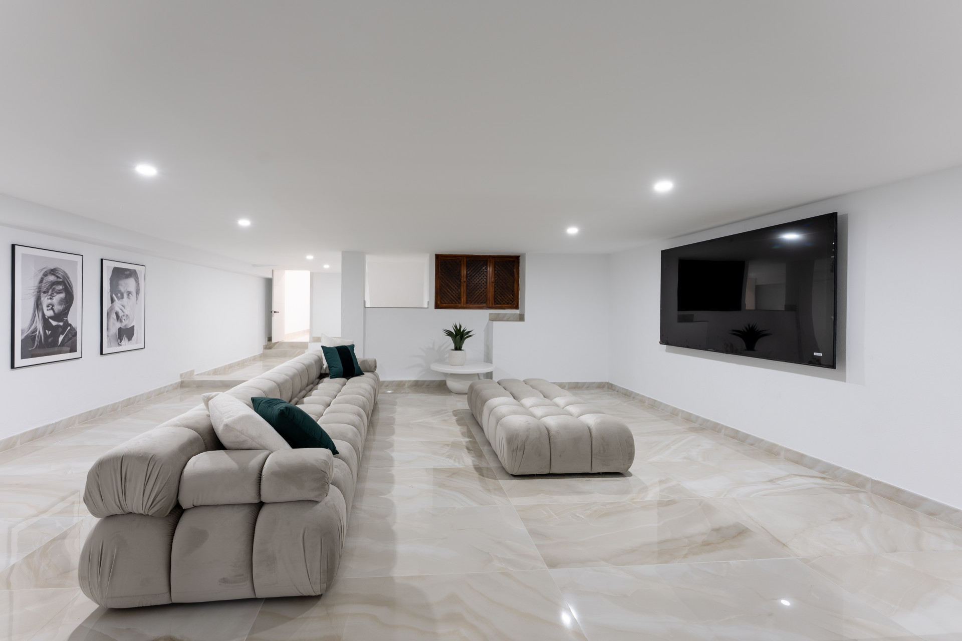 Another view of the media room with a long beige sofa, light beige ottoman, and wall-mounted TV.