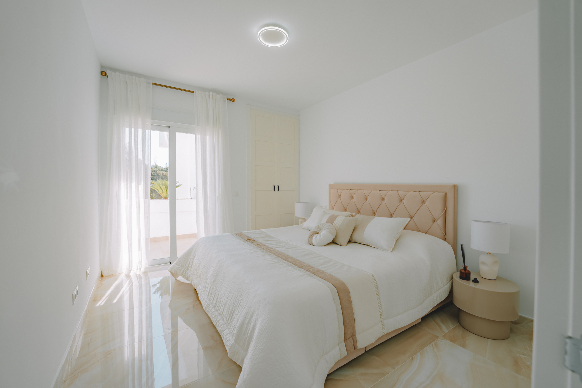 Bedroom with a bed, light wood headboard, and bedside tables. A large glass door with sheer curtains opens to a balcony.