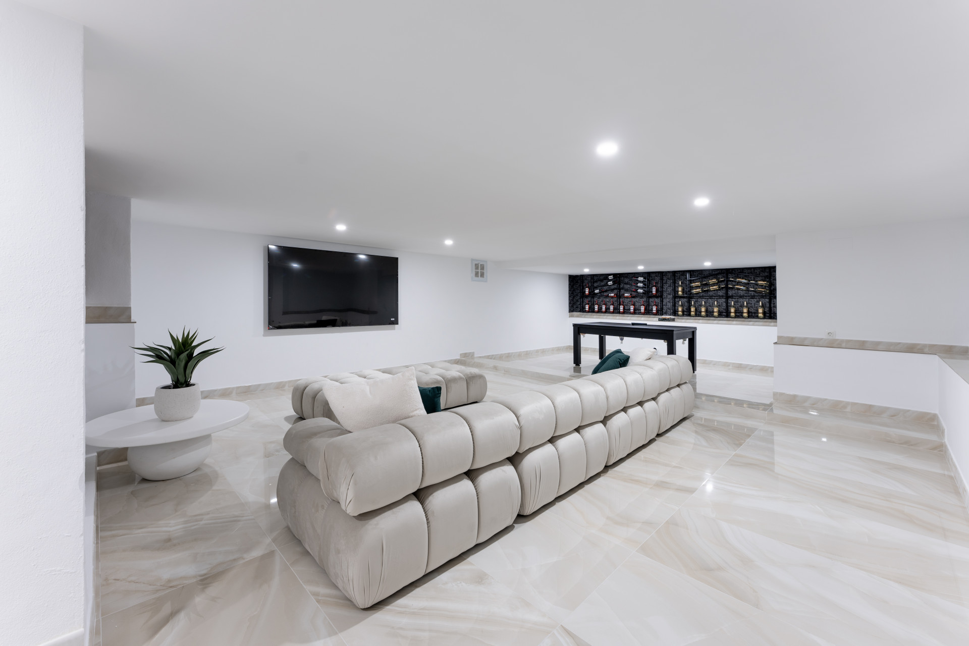 Spacious media room featuring a large TV, long beige sofa, and a black pool table with wine racks.