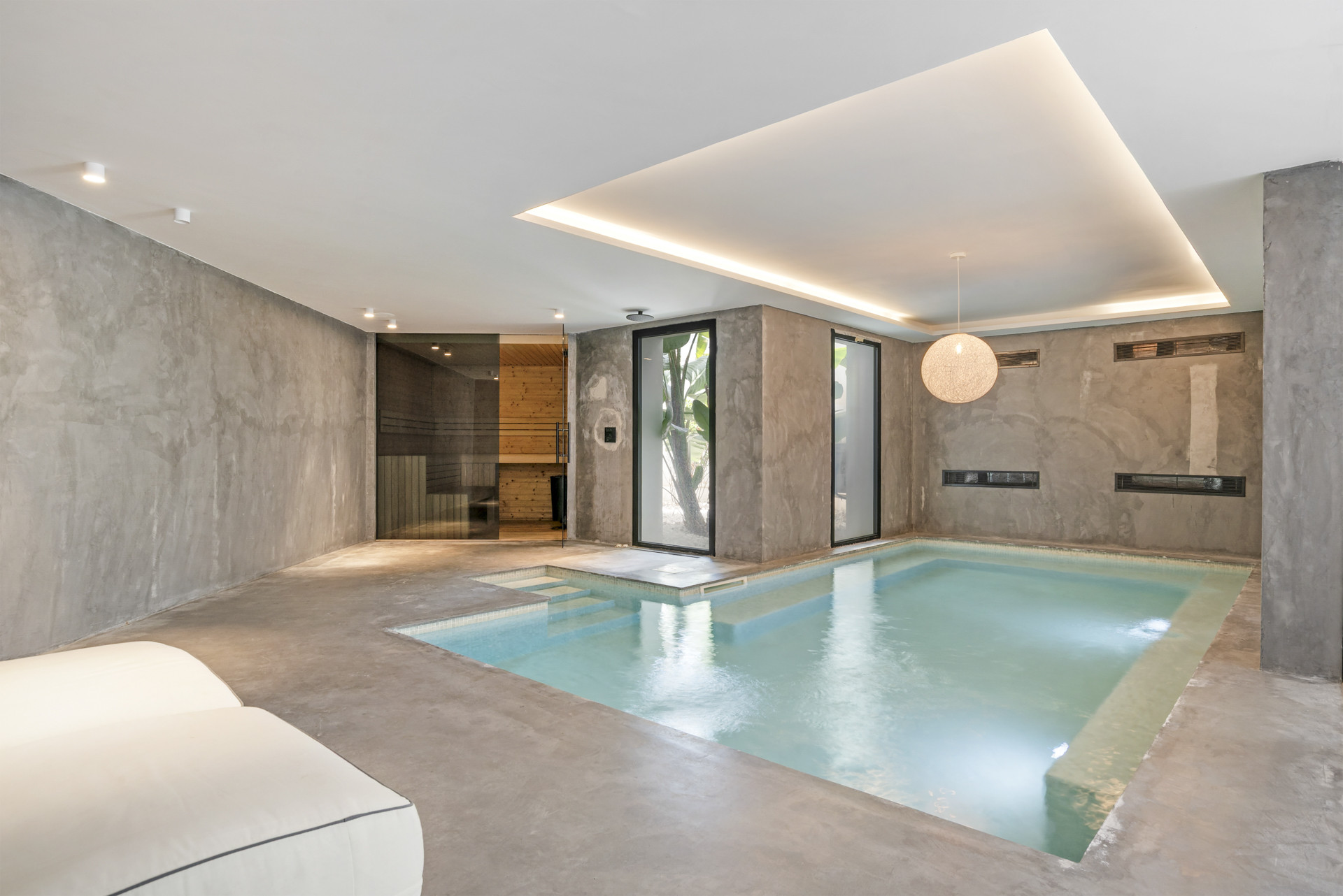 Indoor pool area with soft lighting, concrete walls, and adjacent sauna room visible beyond.