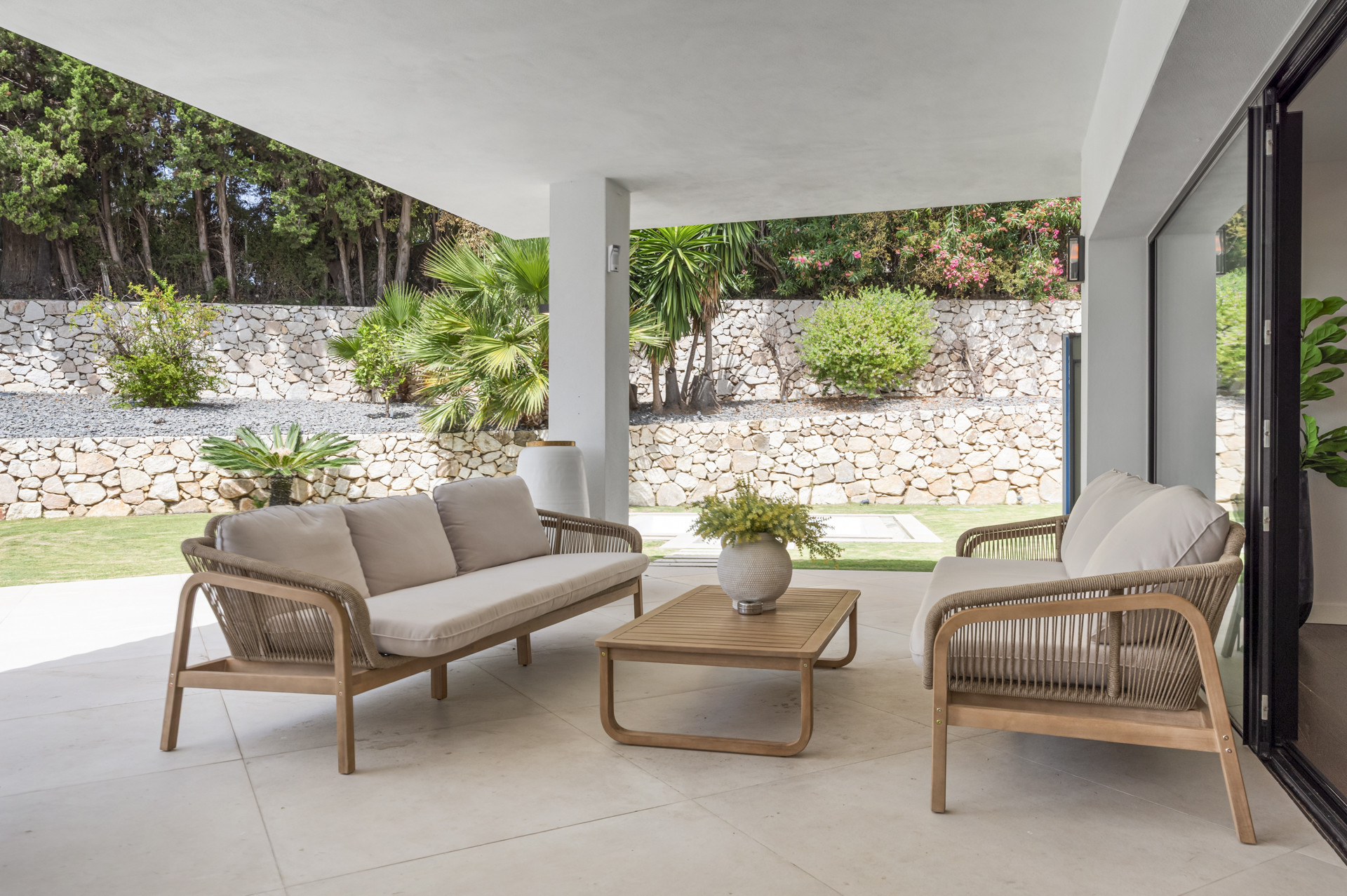 Seating area with rattan furniture and garden view featuring stone walls.
