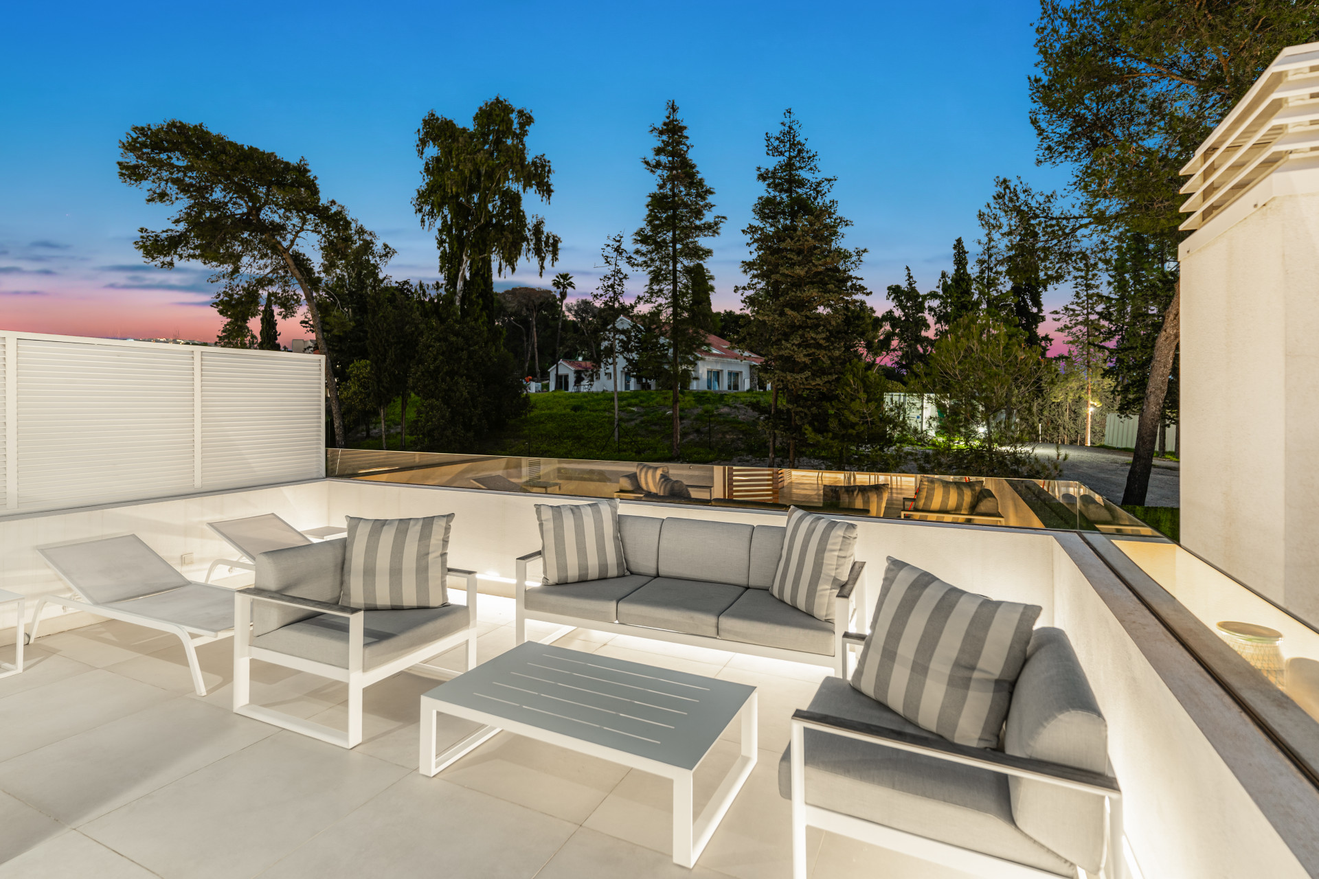 Stylish balcony area with seating, overlooking a green space and trees at dusk.