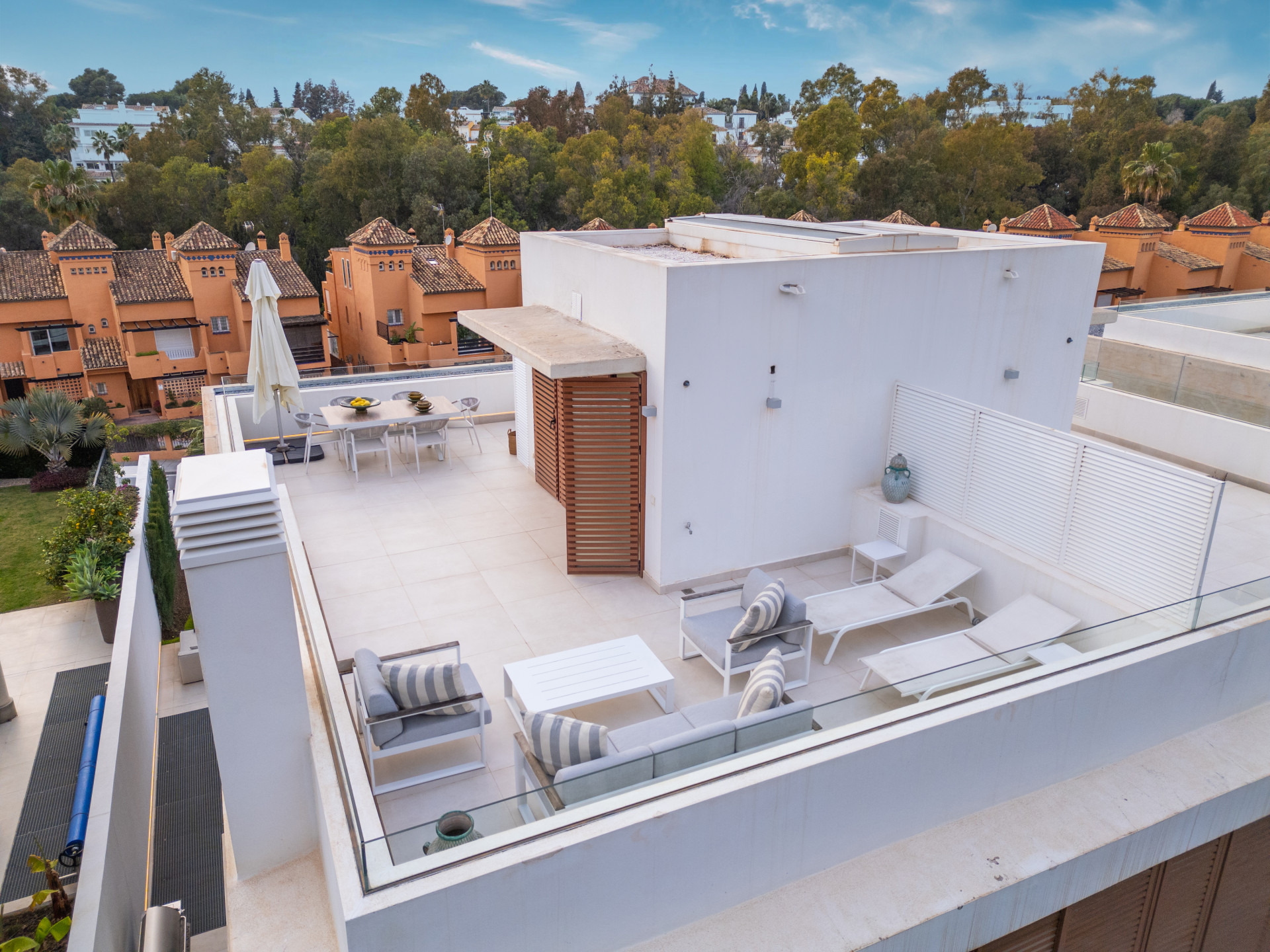 A rooftop terrace with sun loungers, dining area, and views of surrounding trees and buildings.