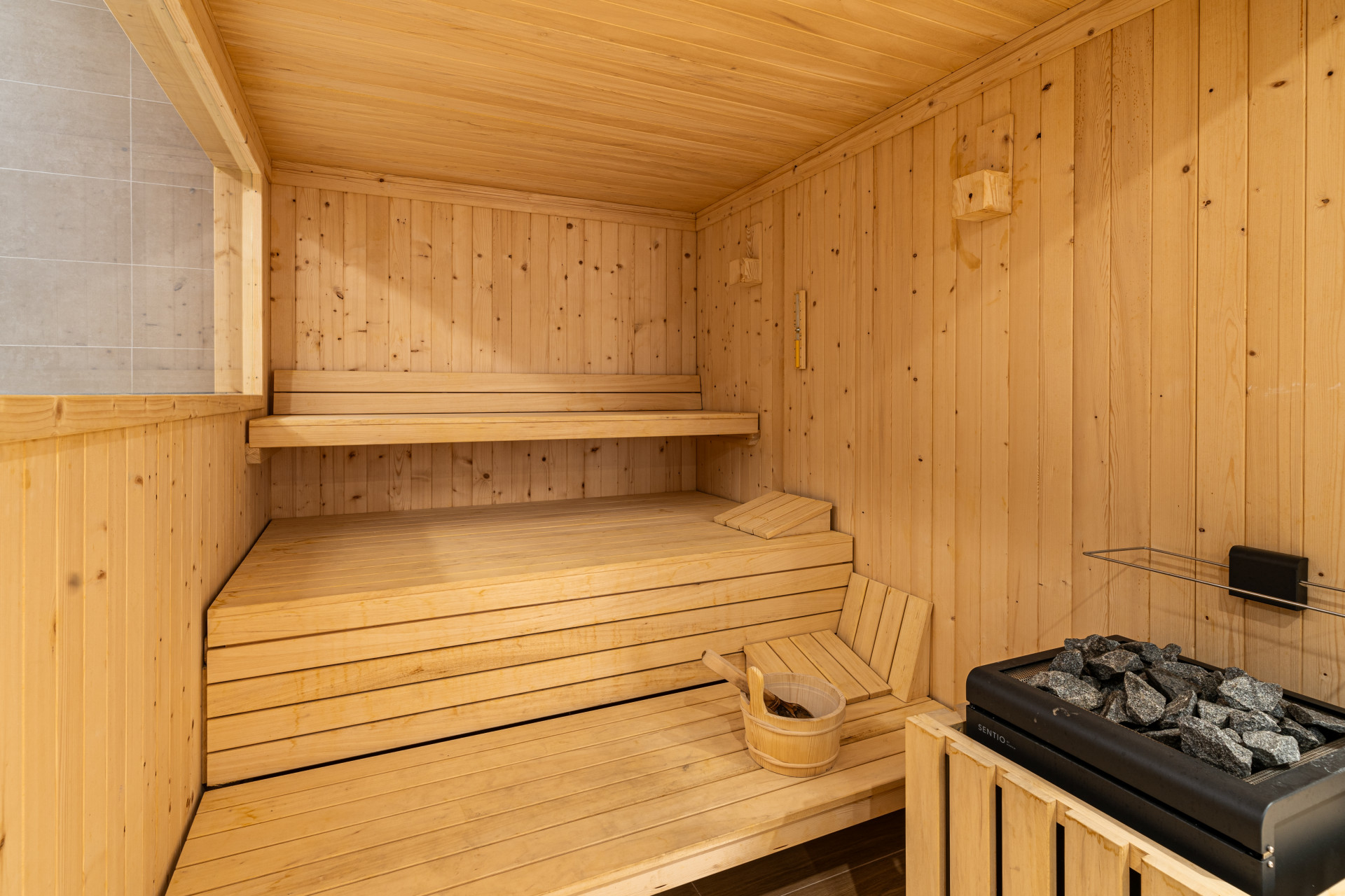 Wood-paneled sauna with benches and a stone heater. A wooden bucket is on the floor.