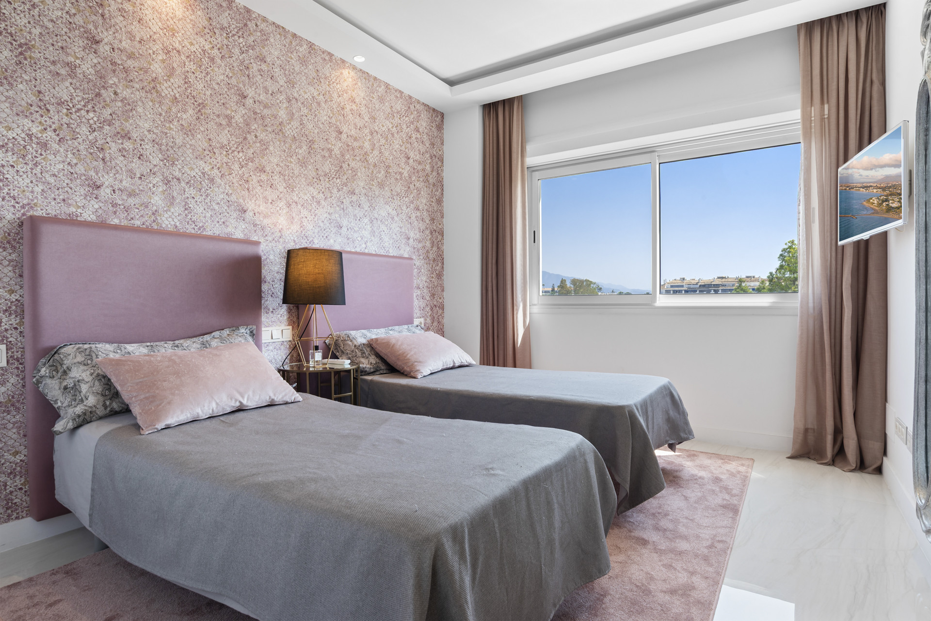 Guest bedroom with two beds, decorative wallpaper, large window, and a wall-mounted TV.