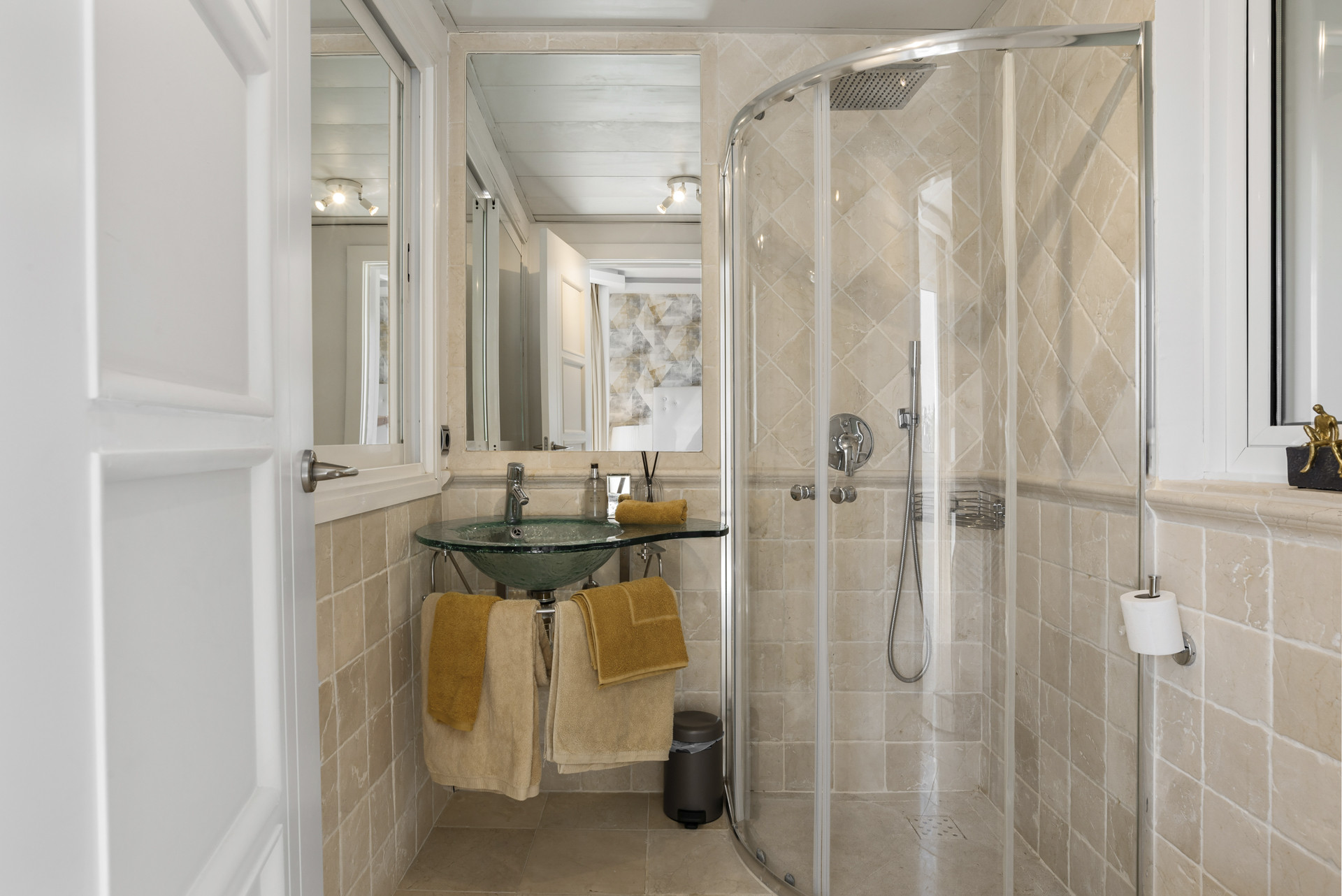 Contemporary bathroom with glass sink, shower, and beige tile walls, showcasing neatly arranged towels.