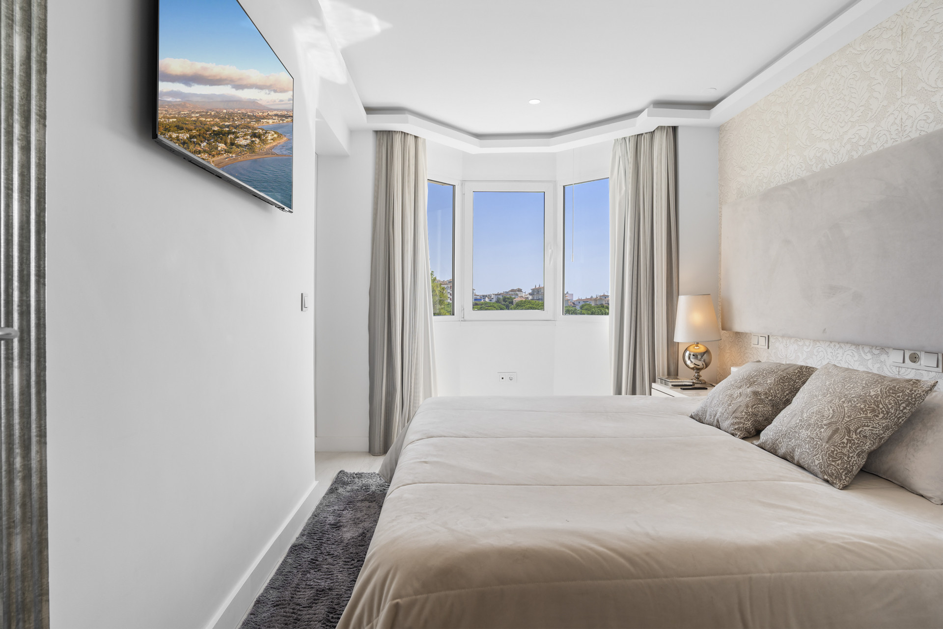 A bright bedroom with a large bed, bay windows, and a TV mounted on the wall.