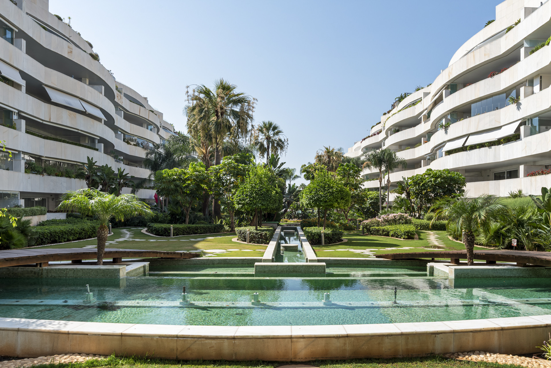 Pathway and water features surrounded by lush palm trees and white buildings.