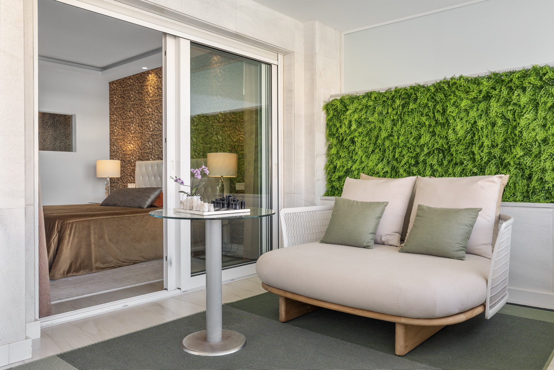 A serene seating area outside a bedroom, featuring a glass table and greenery backdrop with soft cushions.