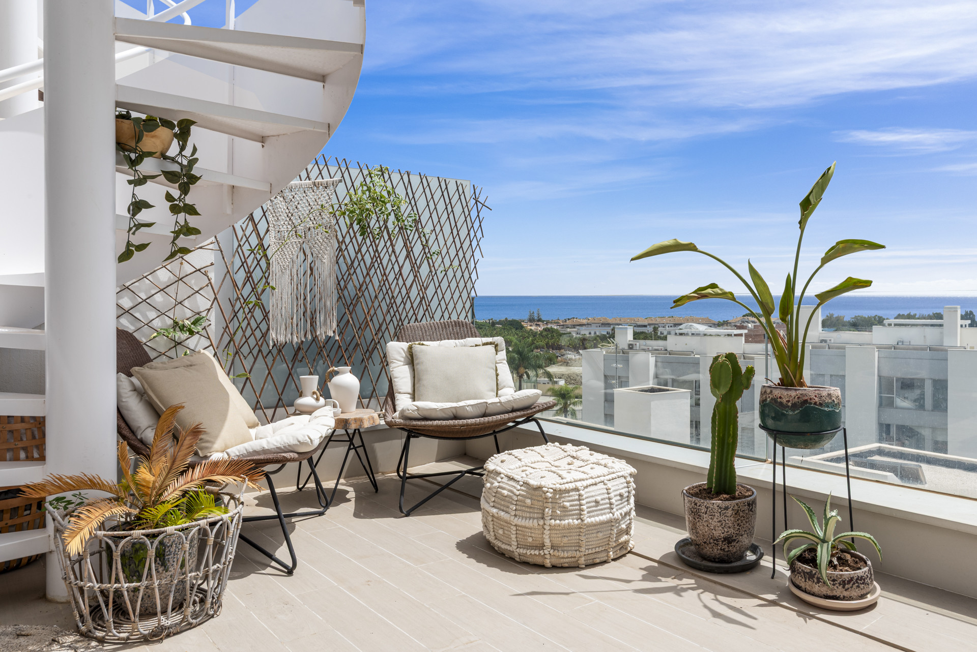 A spiral staircase leads to an upper balcony with seating, potted plants, and distant views of the sea and town.