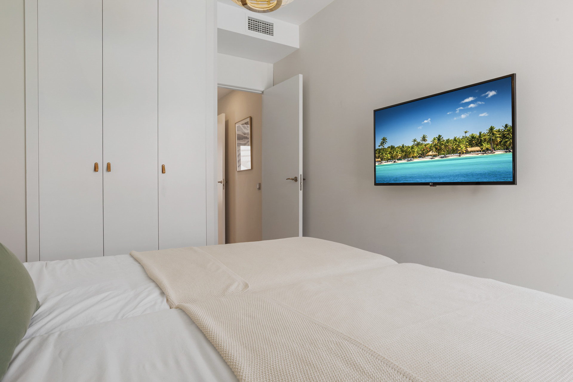 Bedroom with two beds, built-in wardrobes, and a wall-mounted TV displaying a beach scene. An open door is visible.