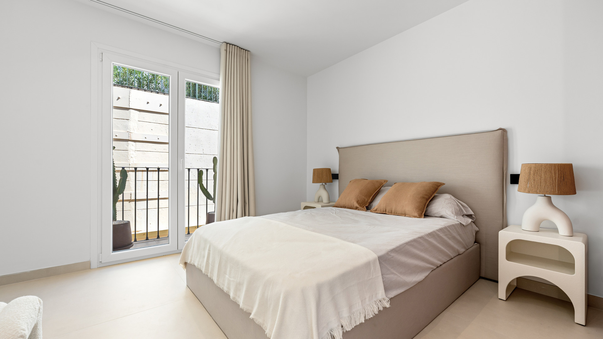 Modern bedroom with a bed, cream-colored cushions, and large windows overlooking a balcony.